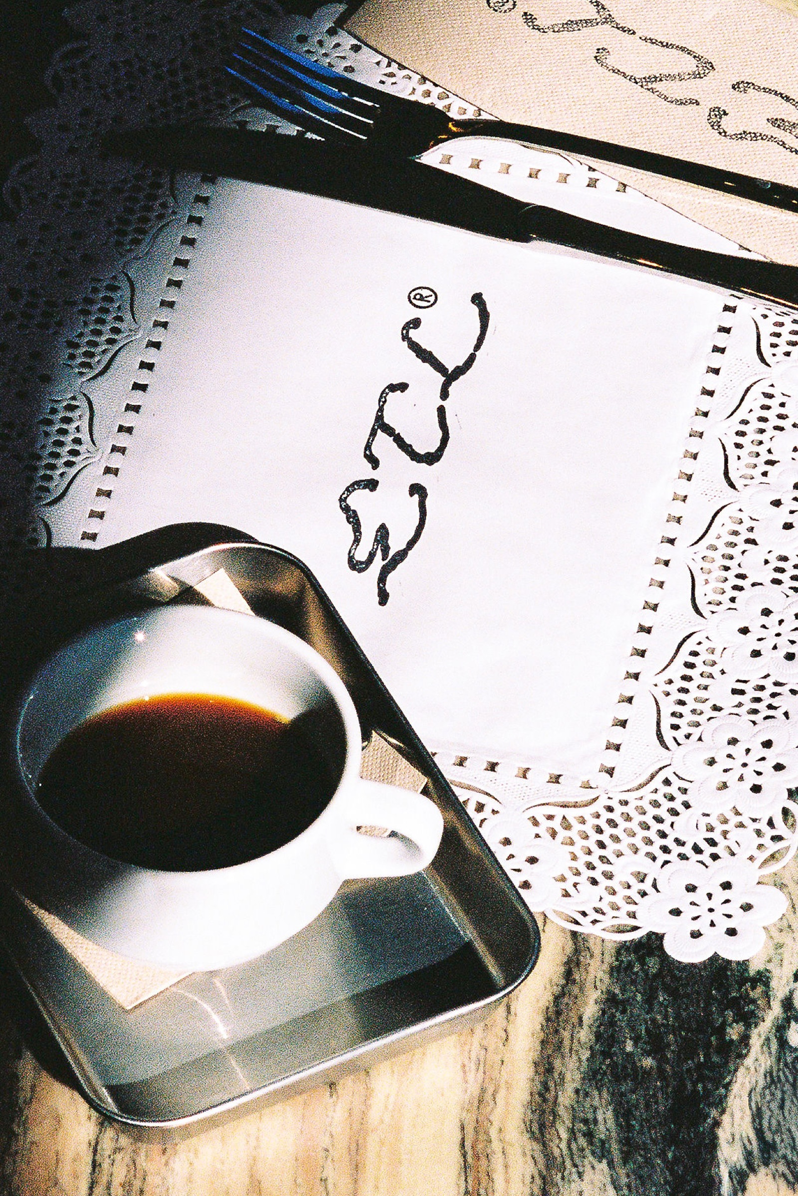 Una taza de espresso sobre una bandeja metálica, junto a una servilleta de papel calado con la firma cursiva ETL escrita a mano y un par de cubiertos plateados.